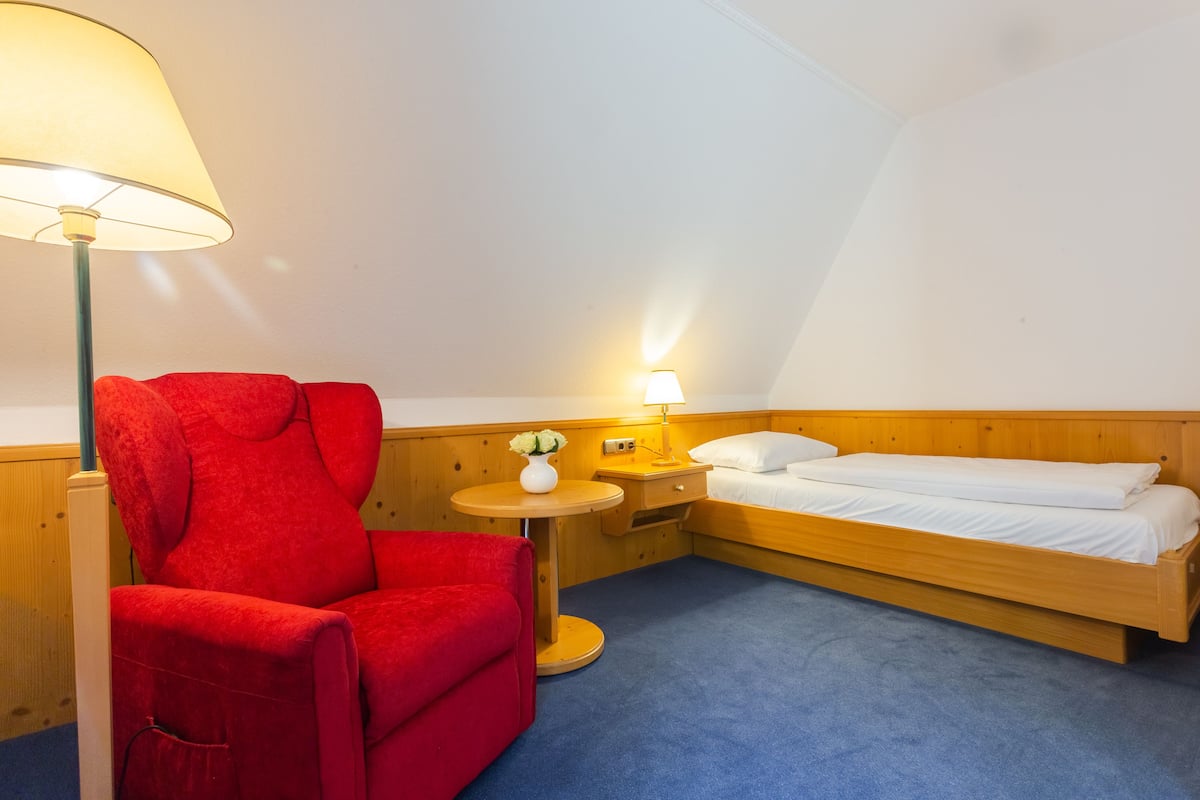 A small, cozy room with a single bed, a red armchair, a small table with a lamp, and a nightstand.