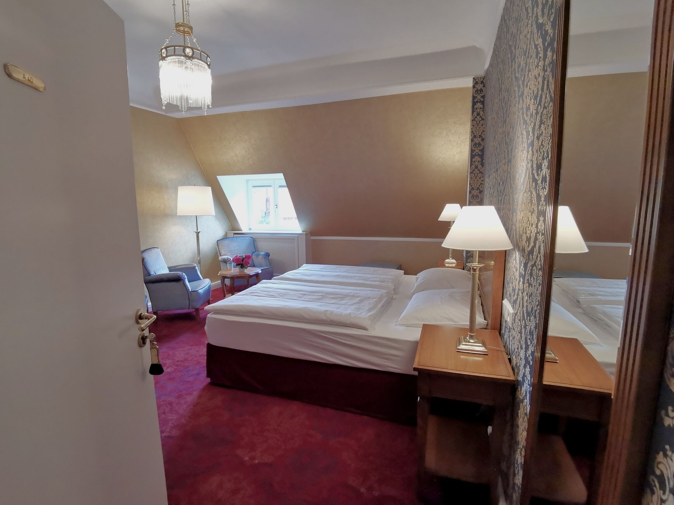 Une chambre d’hôtel avec un grand lit, un bureau, une chaise, une lampe et un lustre.