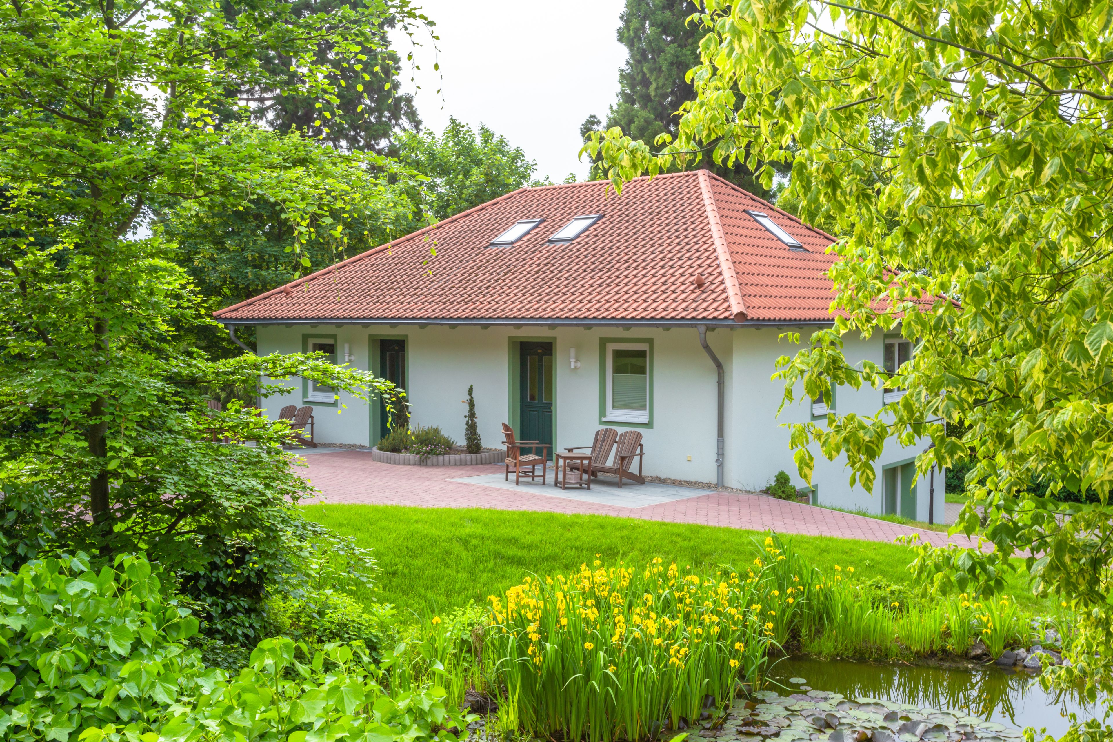 Ein Haus mit rotem Ziegeldach, einer Veranda mit zwei Stühlen und einem kleinen Teich mit gelben Blumen im Vordergrund.