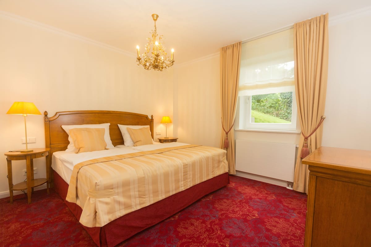 A hotel room with a large bed, two bedside tables, a chandelier, a window with curtains, and a red carpet.