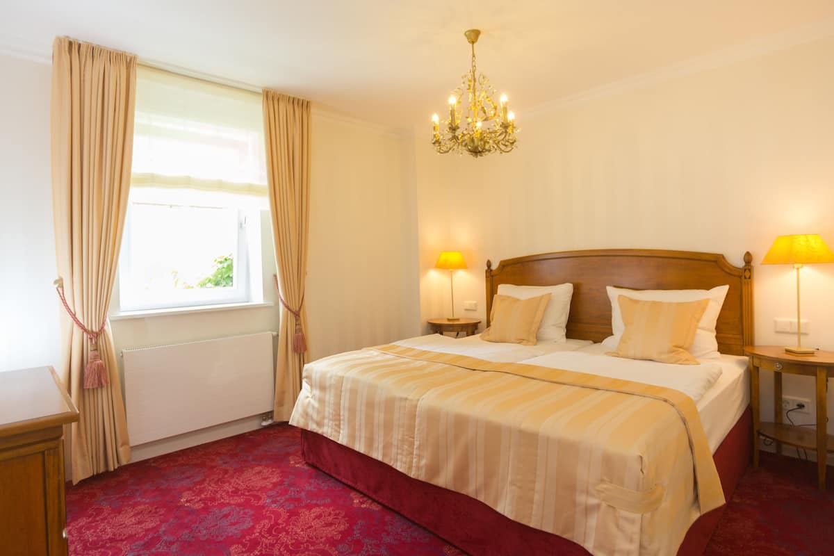 A hotel room with a large bed, two bedside lamps, a chandelier, and a window with curtains.