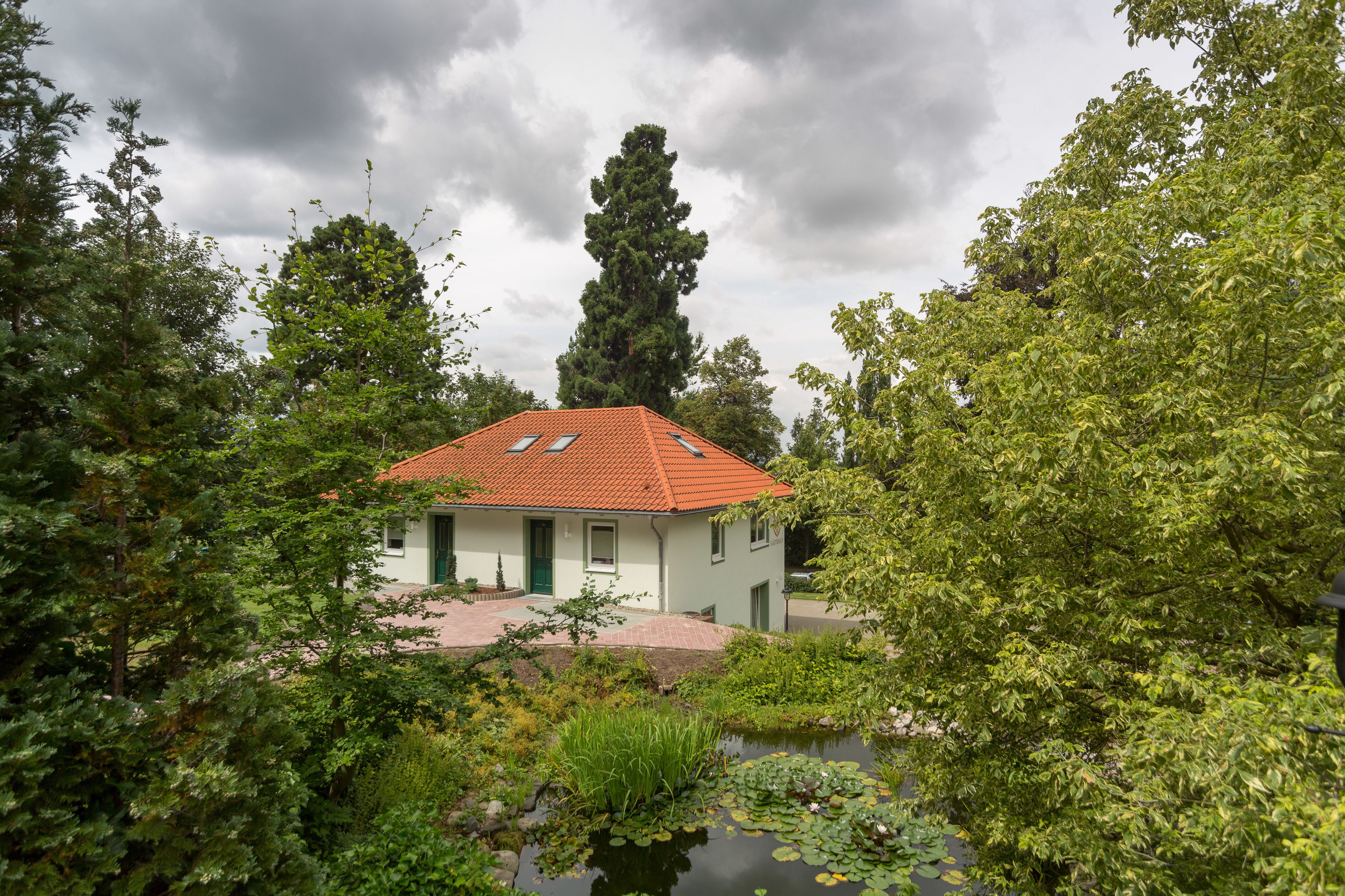 Een huis met een rode pannendak, omgeven door weelderige begroeiing en een vijver.