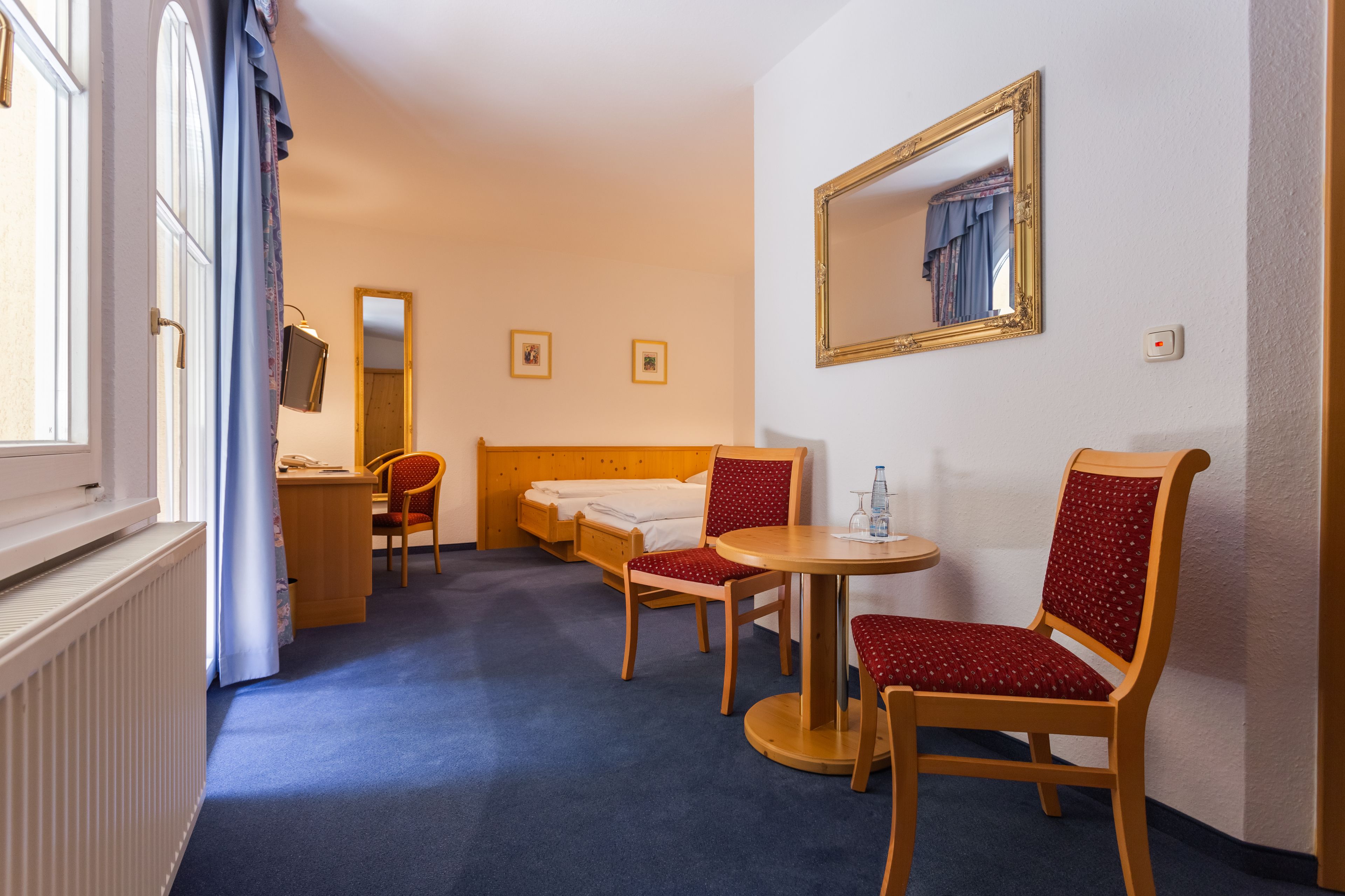 A hotel room with a bed, two chairs, a small table, a mirror, and a window with curtains.