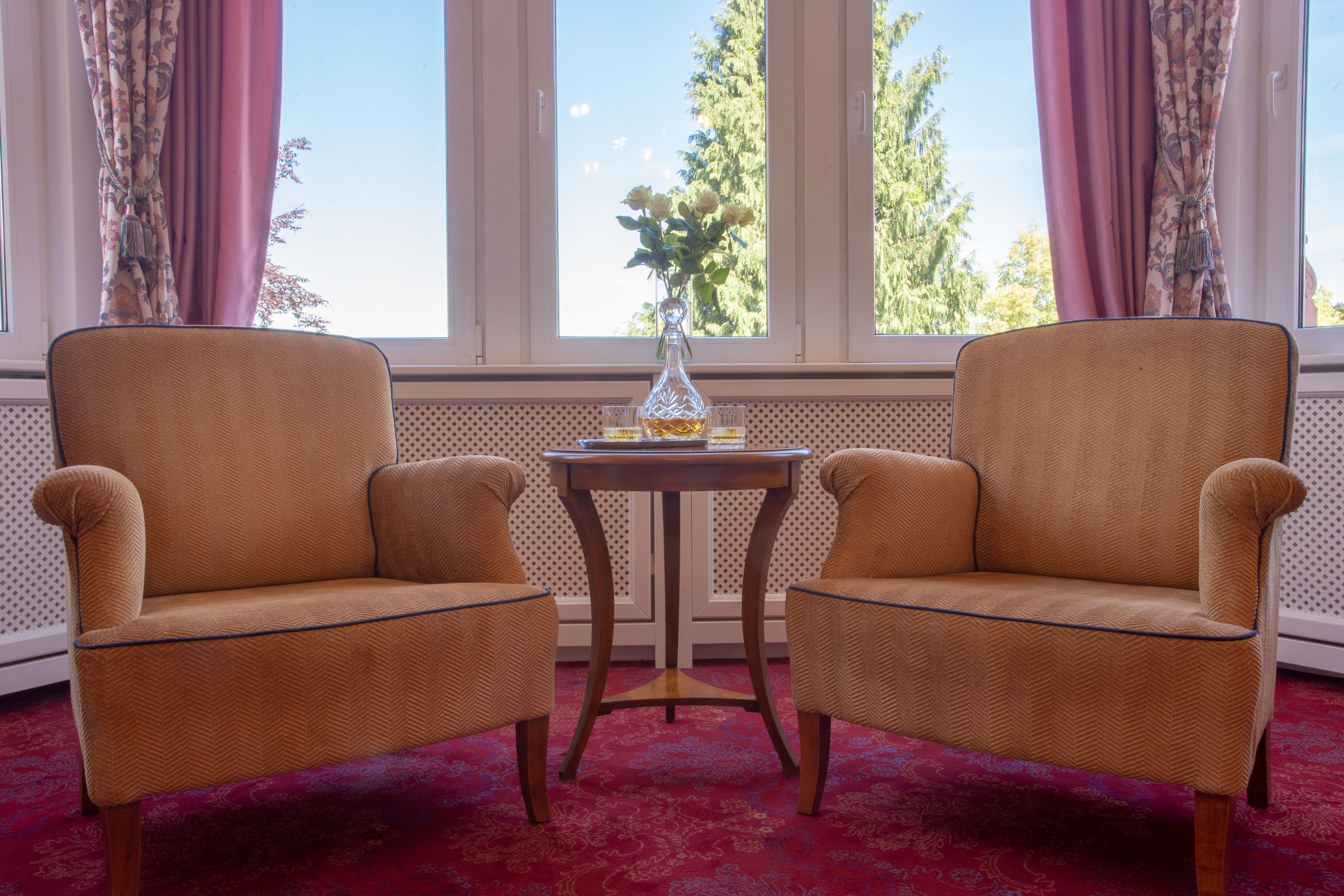 Two armchairs with a small table between them, a vase with flowers, and a window with curtains.