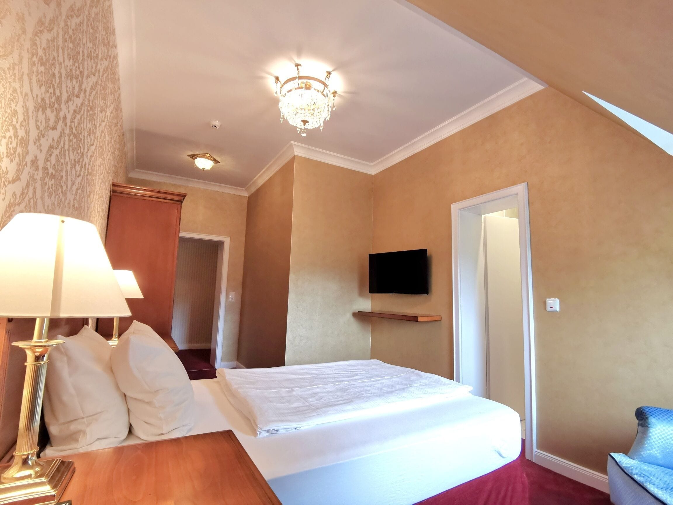Una habitación de hotel con una cama, un televisor montado en la pared, una lámpara y una mesita de noche de madera.