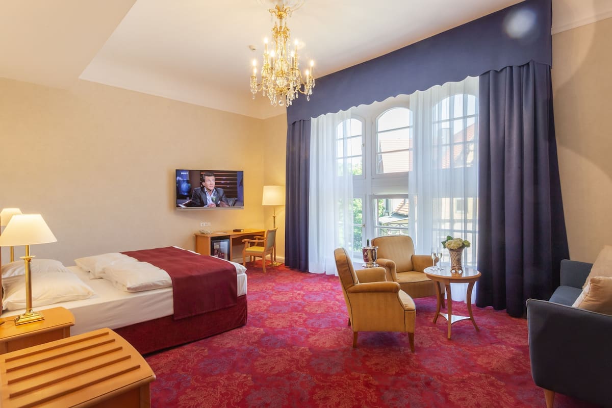 A hotel room with a bed, a desk, a television, a chandelier, and a view through large windows.