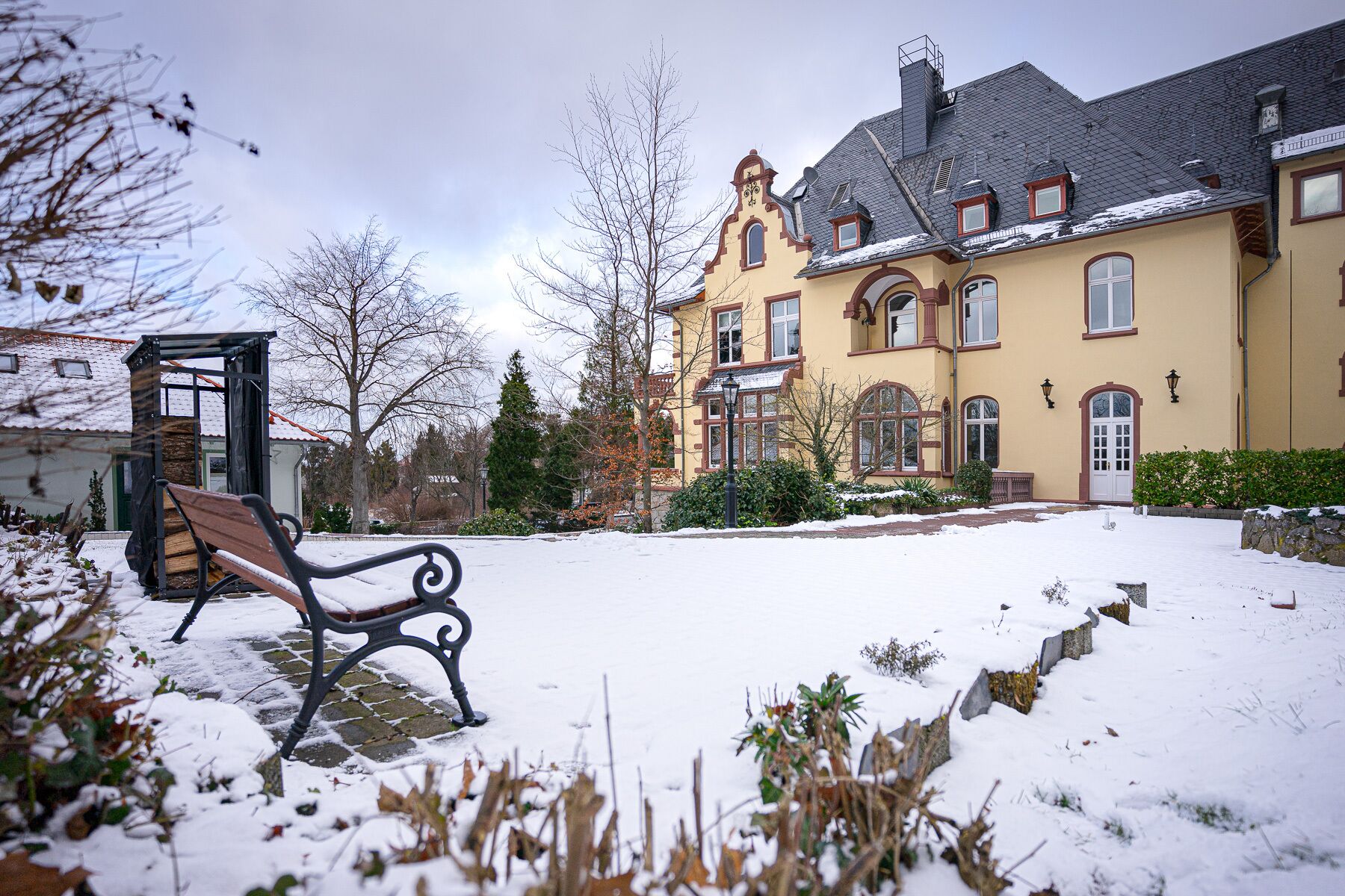 A snow-covered courtyard with a bench, a yellow building with multiple windows, and a small structure with a red roof.