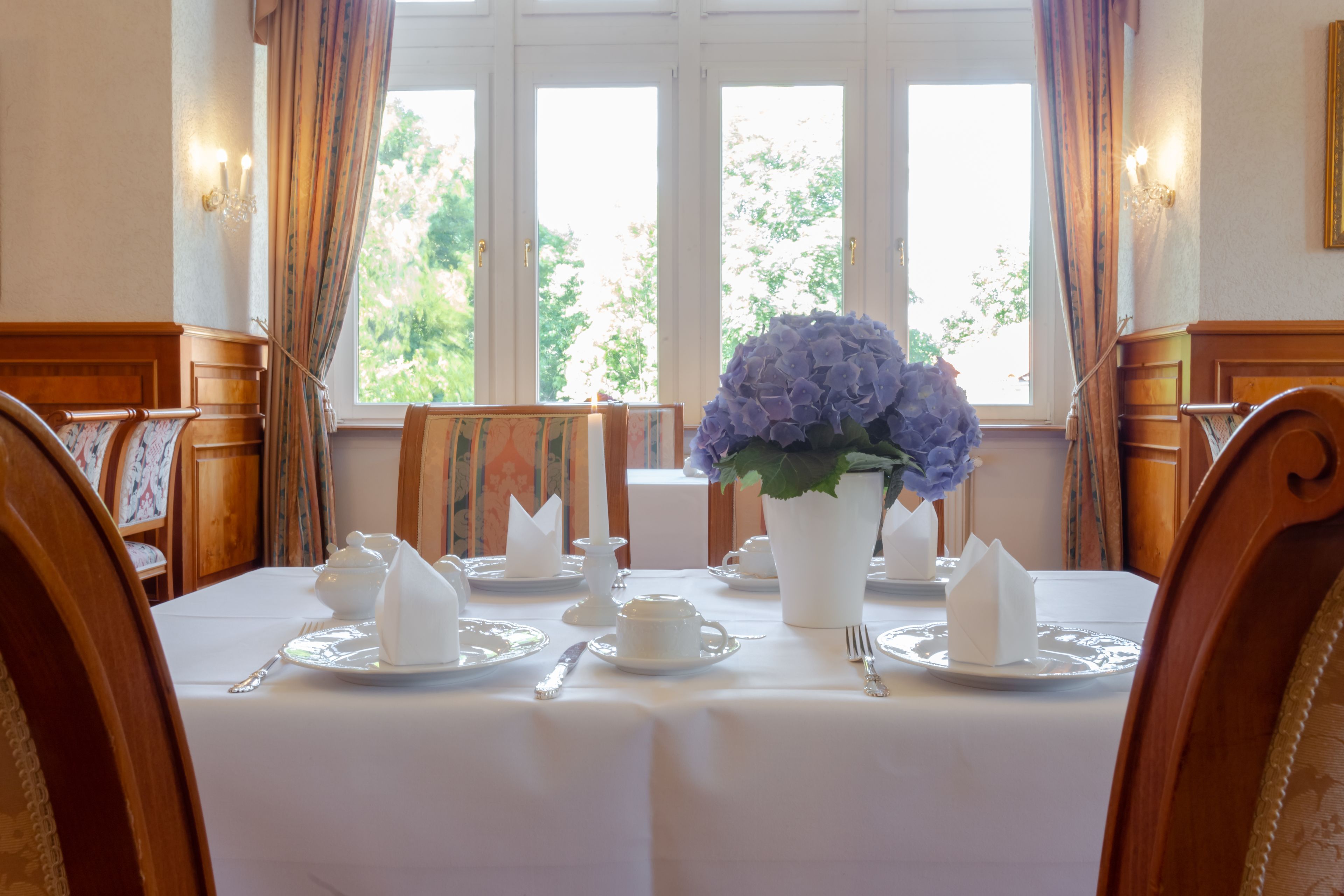 A dining room with a table set for four, featuring white tablecloths, silverware, and a vase of flowers.