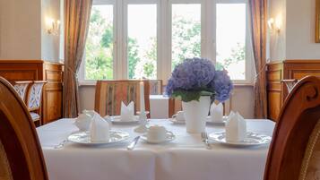 A dining room with a table set for four, featuring white tablecloths, silverware, and a vase of flowers.
