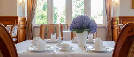 A dining room with a table set for four, featuring white tablecloths, silverware, and a vase of flowers.