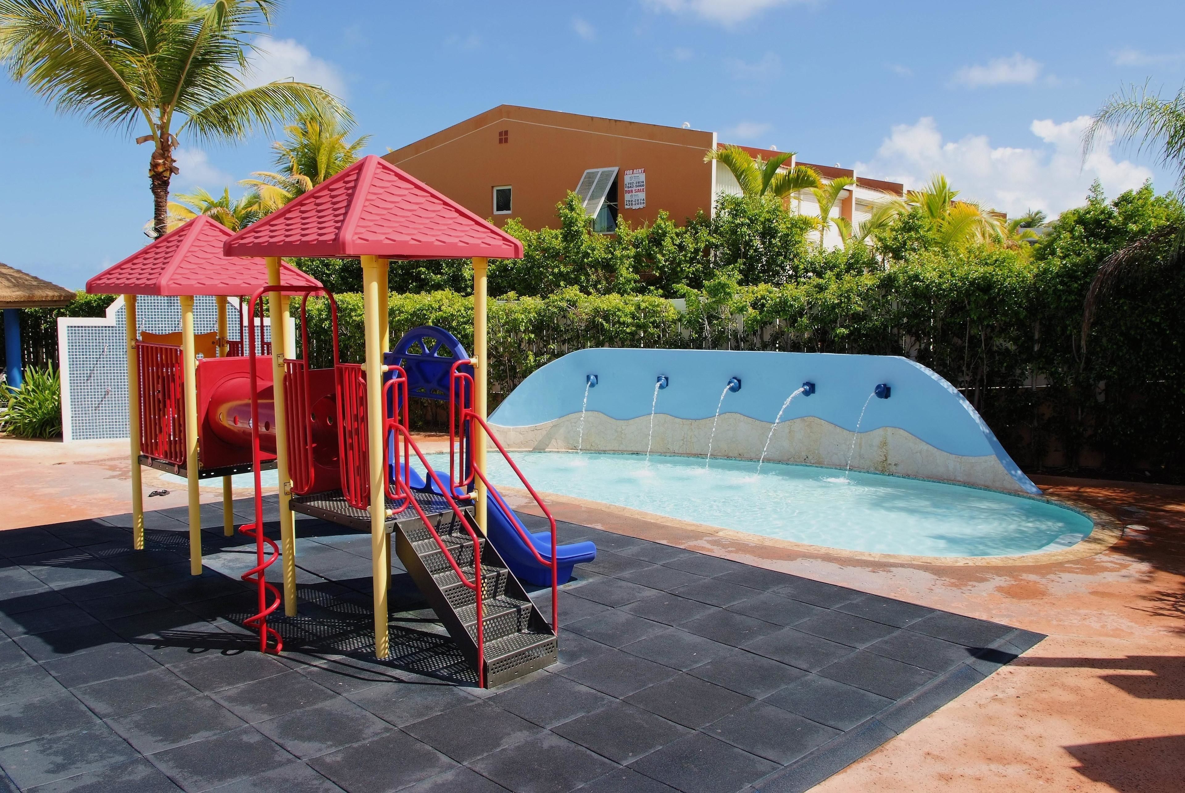 Un parque infantil con tobogán y zona de escalada, una piscina con diseño de olas y un edificio al fondo.