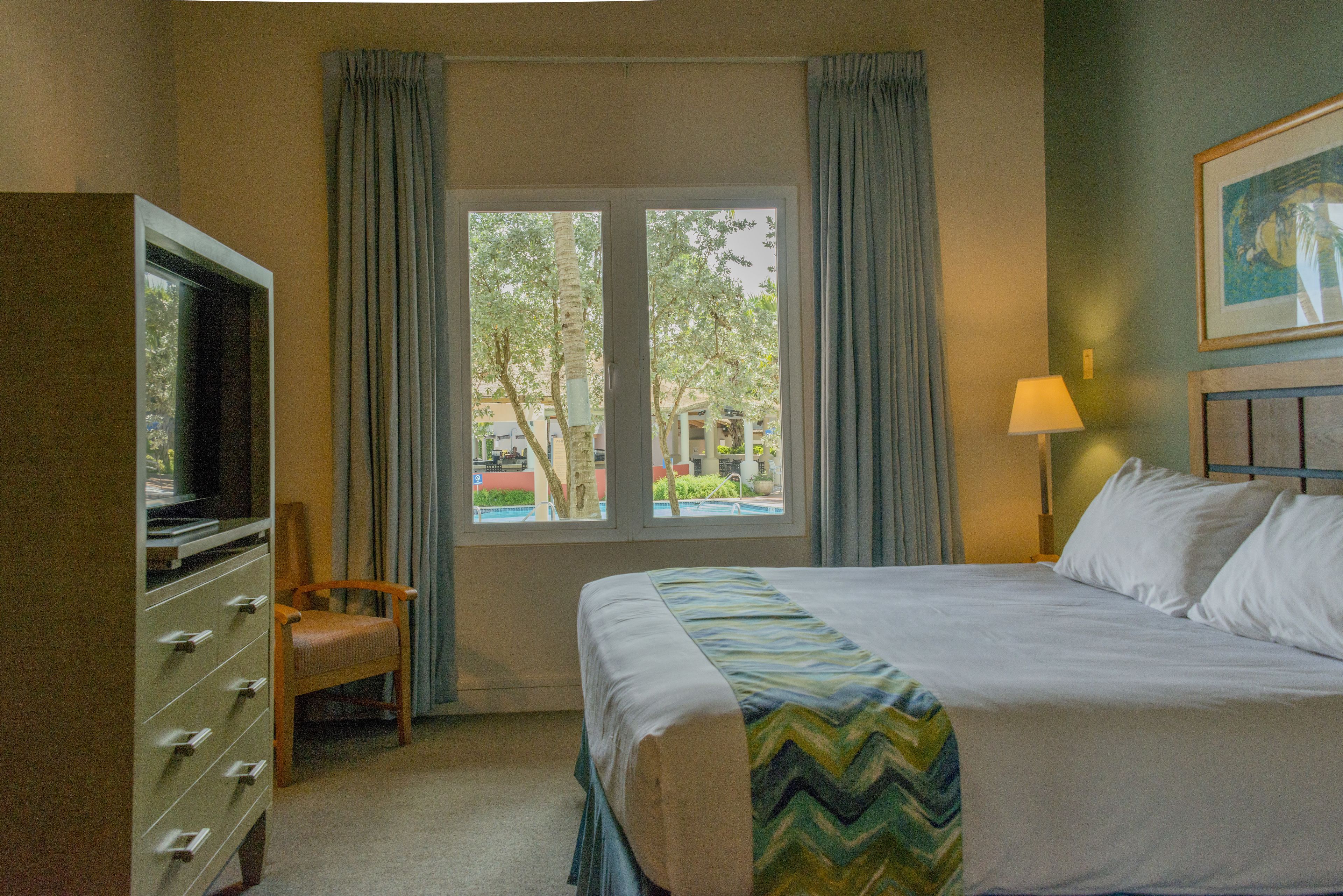 Una habitación de hotel con cama, televisión, silla, ventana con cortinas y lámpara.