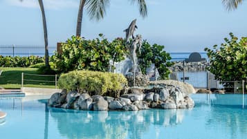 Una piscina azul cristalina con una formación rocosa, palmeras y una torre de salvavidas al fondo.