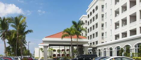 A hotel entrance with a red-tiled roof, palm trees, and a parking lot with various cars.