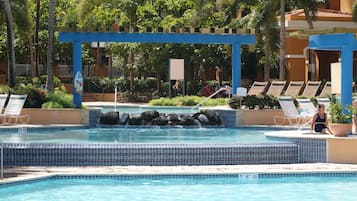 Ett poolområde med solstolar, en blå pergola och ett litet vattenfall.