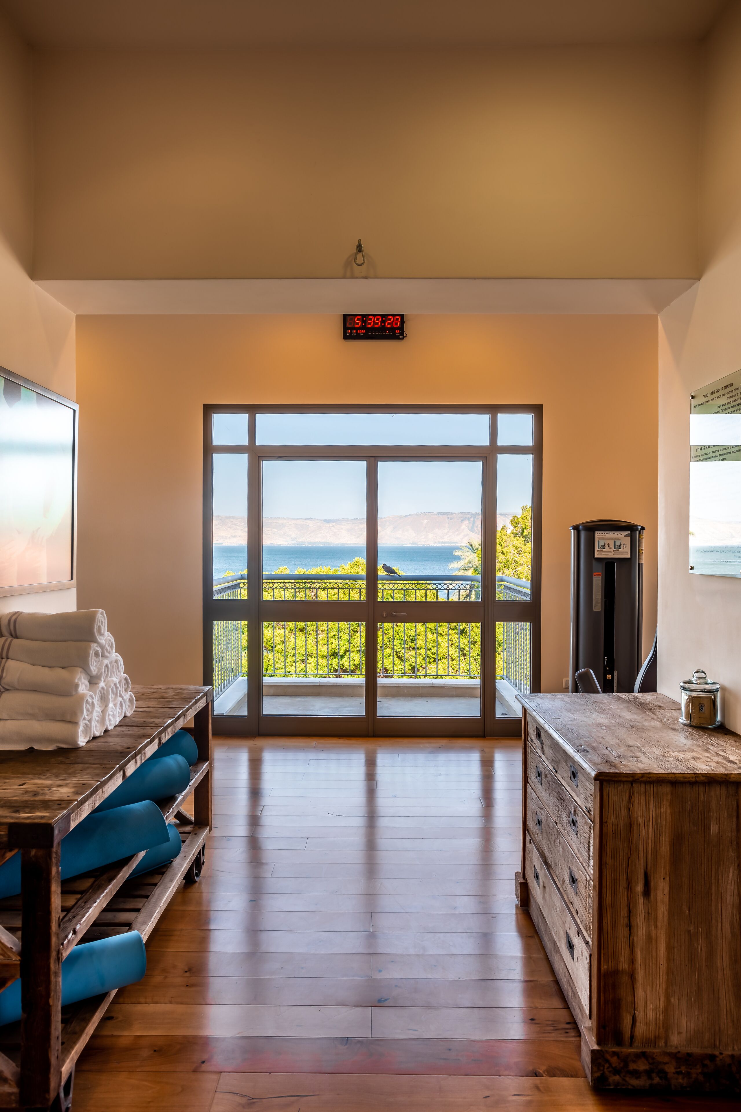A spacious room with a large sliding glass door leading to a balcony with a view of the sea and hills. There is a wooden bench with towels, a small table, and a cabinet with a water dispenser.