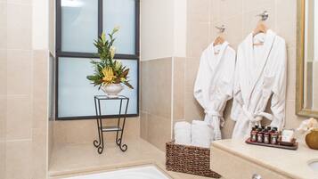 A modern bathroom with a bathtub, a window with frosted glass, a small plant on a metal stand, and two white robes hanging on hooks.