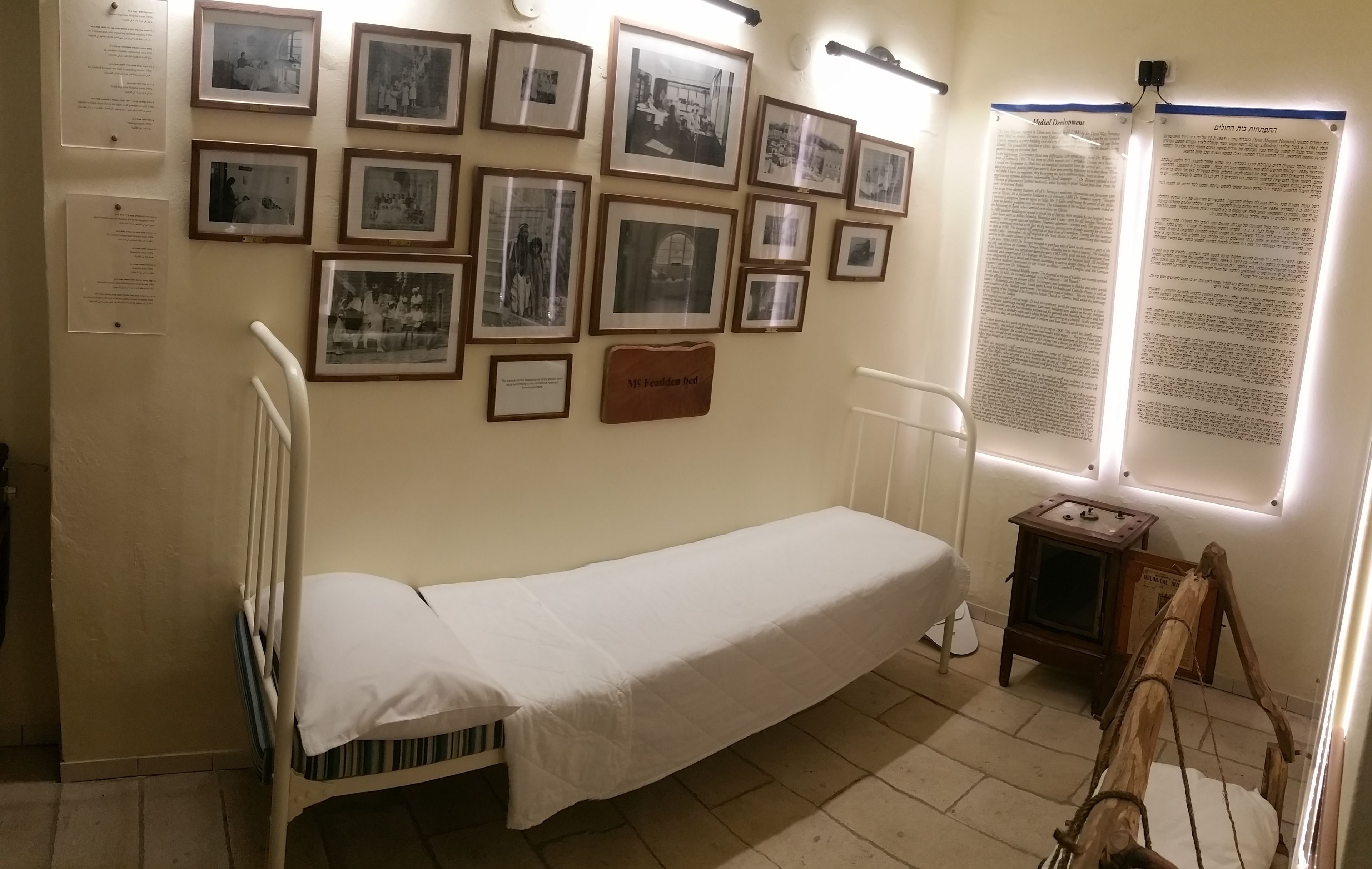 Ein Zimmer mit einem Einzelbett, einem hölzernen Kopfteil, einem kleinen Nachttisch und einer Wand mit gerahmten Fotografien und Infotafeln.