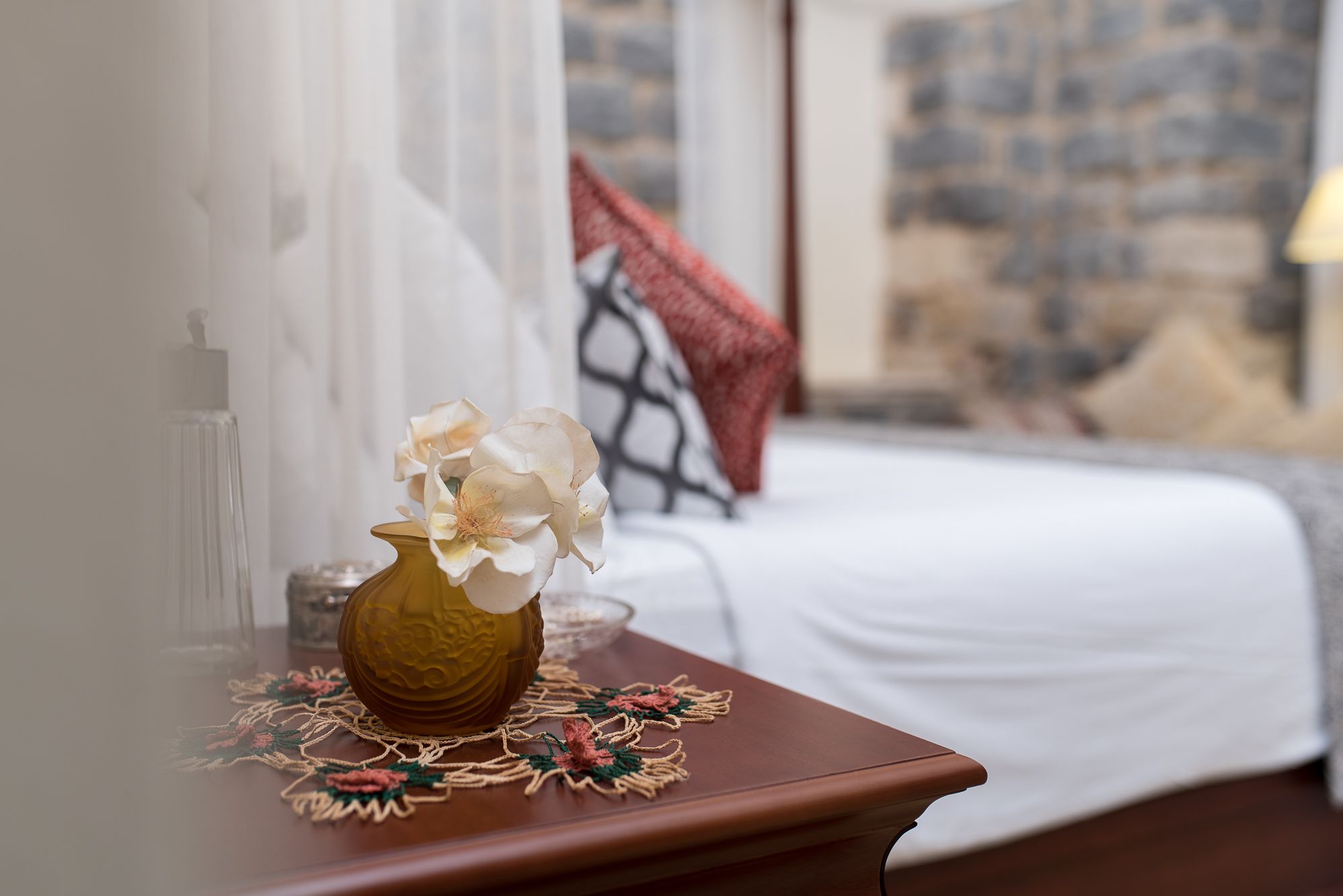 Un dormitorio con una cama, una mesita de noche con un florero y vista a un muro de piedra.