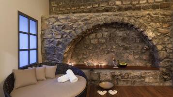 Una chimenea de piedra con estante empotrado, un sillón de mimbre con cojines y una mesa de madera con un cuenco y objetos con forma de corazón.