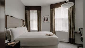Habitación de hotel con una cama grande, una mesita de noche con lámpara, un escritorio con silla y una ventana con cortinas.