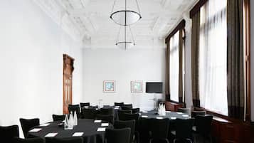Ein Konferenzraum mit hoher Decke, aufwendigen Stuckverzierungen und großen Fenstern mit Vorhängen. Es gibt mehrere Tische mit Stühlen, einen Flachbildfernseher und gerahmte Kunstwerke an den Wänden.