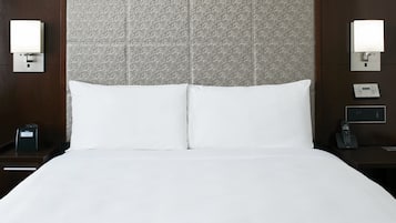 Habitación de hotel con una cama grande, dos mesitas de noche, un reloj de pared, un termostato y dos lámparas fijadas a la pared.