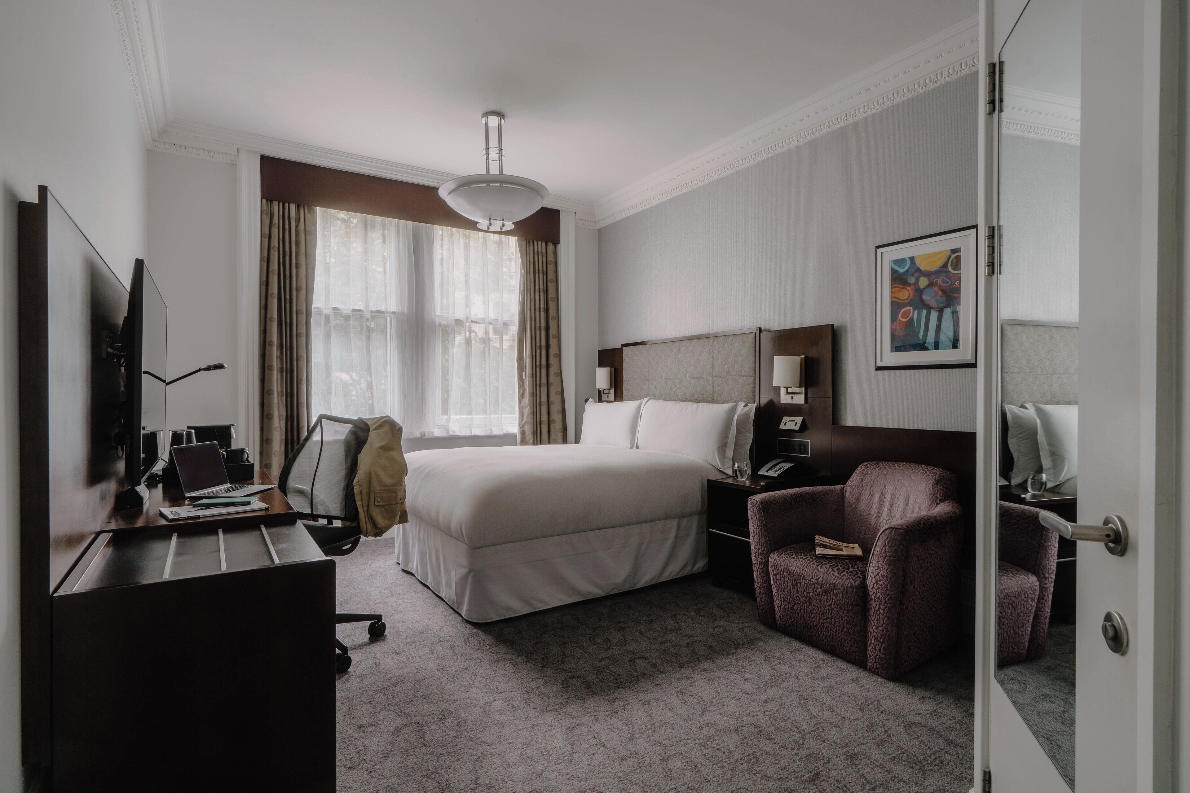 Habitación de hotel con una cama grande, un escritorio con una computadora portátil, una silla, una mesita de noche y un televisor.