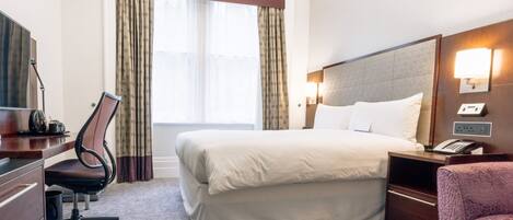 A hotel room with a large bed, a desk with a chair, a desk lamp, a telephone, and a nightstand.