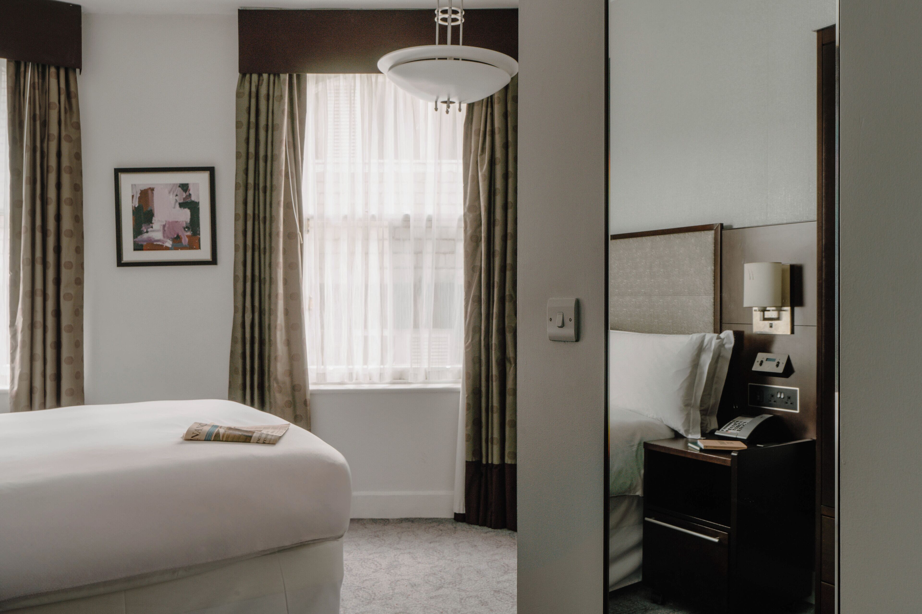 Ein Hotelzimmer mit einem Bett, einem Nachttisch mit Lampe, einem Fenster mit Vorhängen und einem gerahmten Bild an der Wand.
