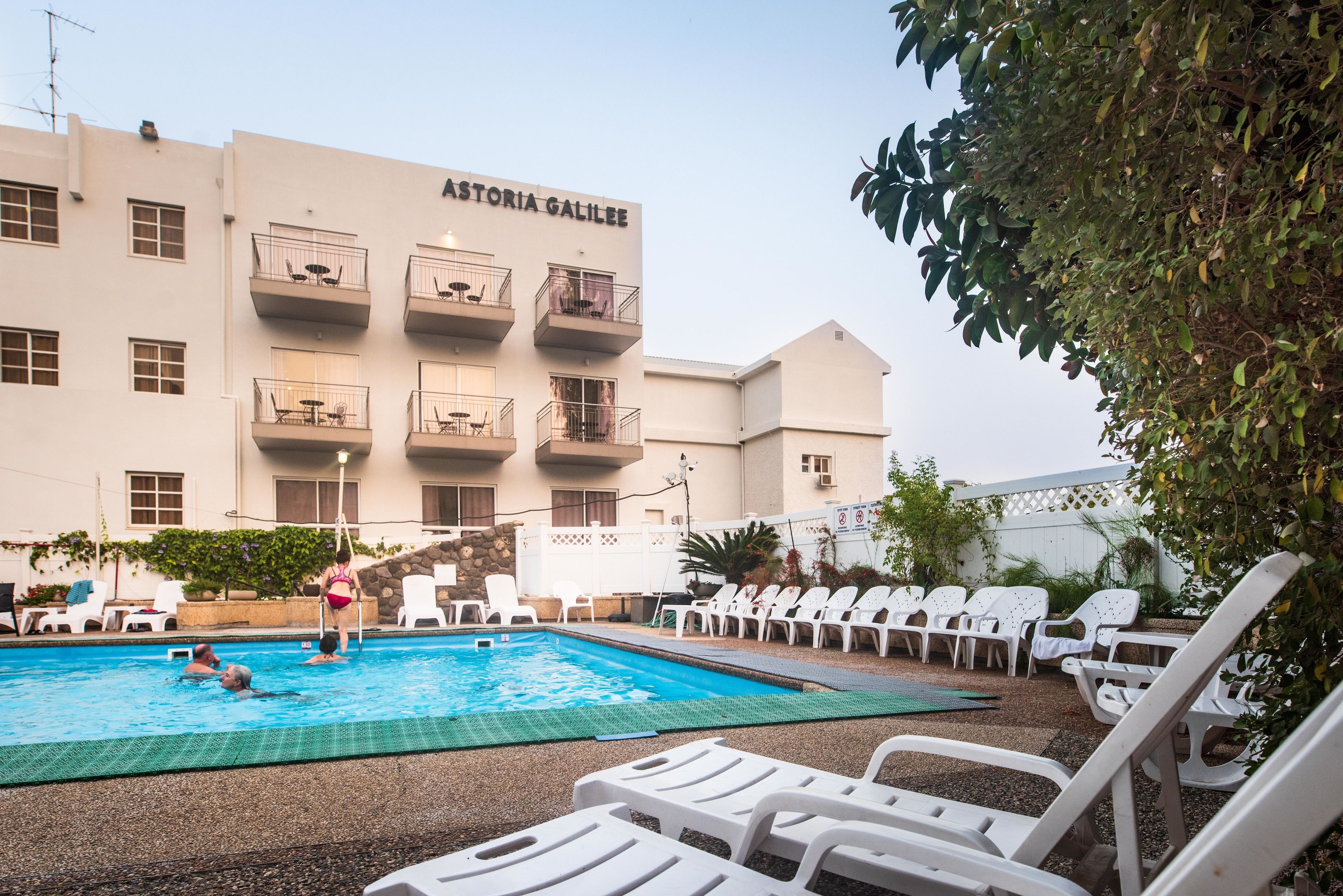 Zona de piscina con sillones de descanso y un edificio con la inscripción 'ASTORIA GALILEE' al fondo.