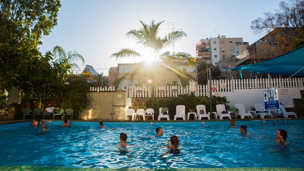 Une piscine avec des personnes dans l’eau, entourée d’une clôture, de palmiers et de bâtiments en arrière-plan.