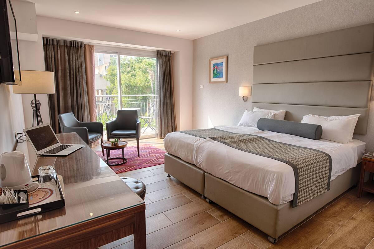 Une chambre d’hôtel avec un lit, un bureau avec un ordinateur portable, une chaise et un balcon avec des rideaux.