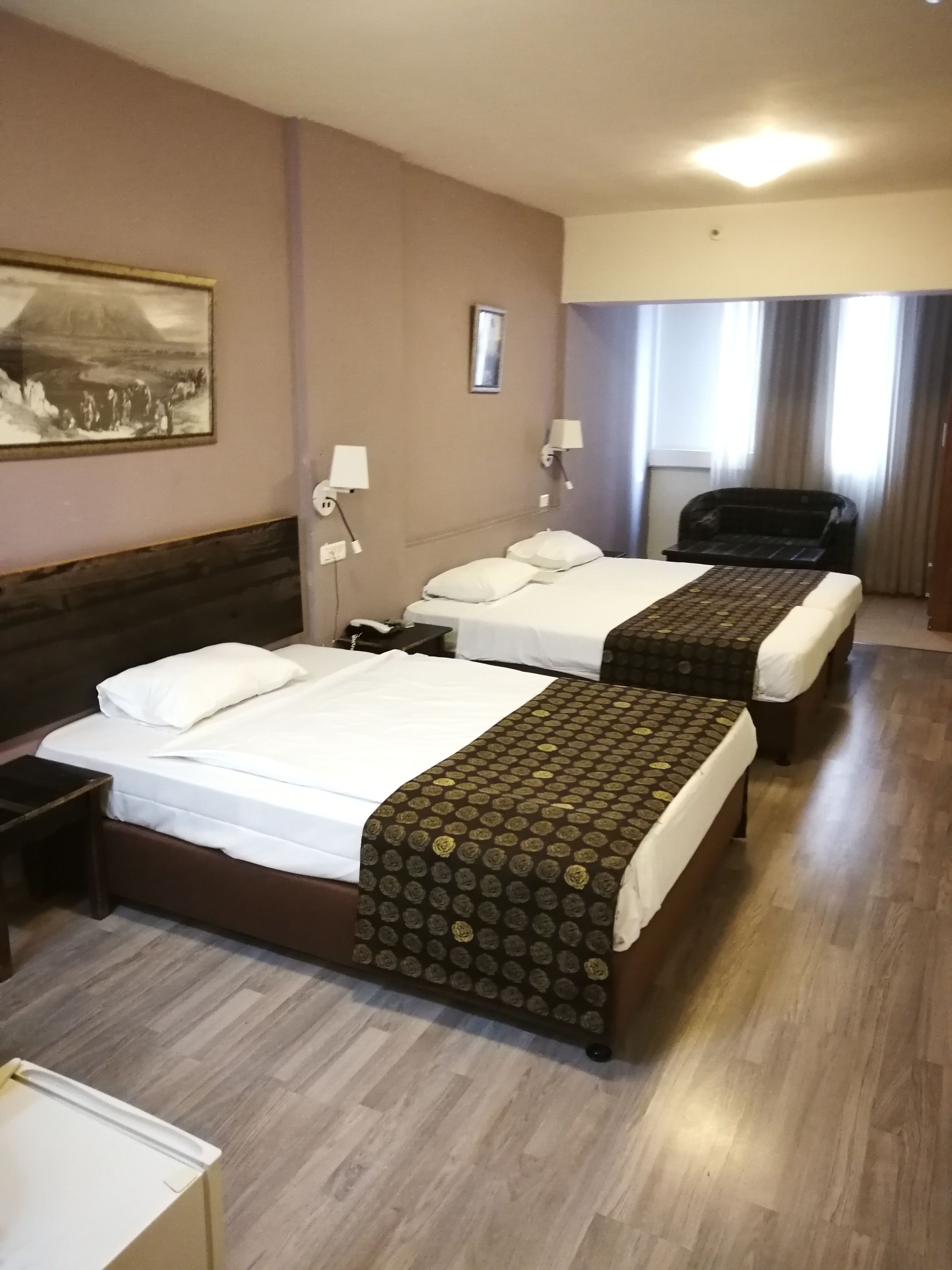 A hotel room with two single beds, a desk, a chair, and a large window with curtains.