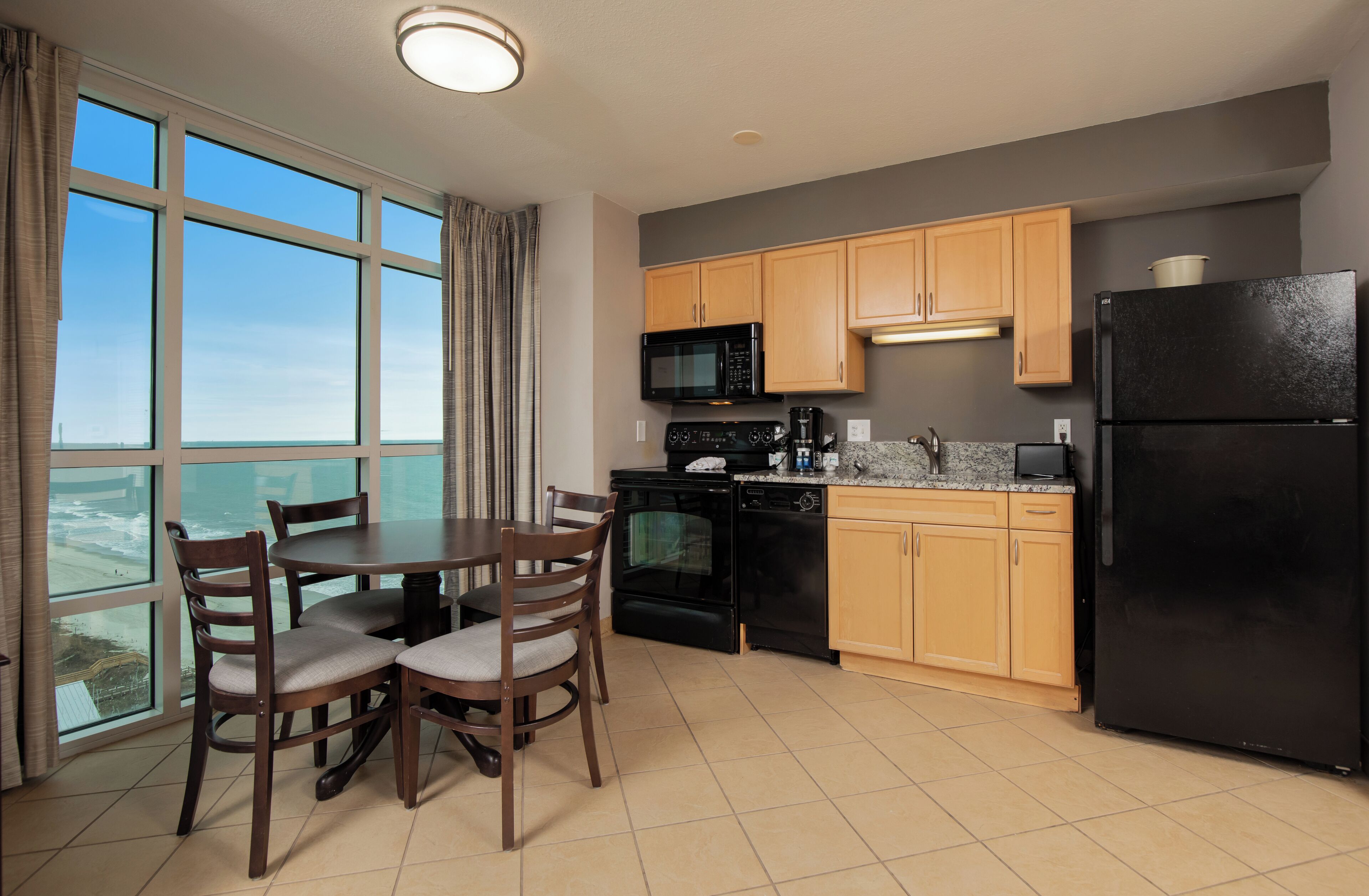 A modern kitchen with wooden cabinets, a black refrigerator, a microwave, and a coffee maker. There is a dining table with chairs and a large window offering a view.