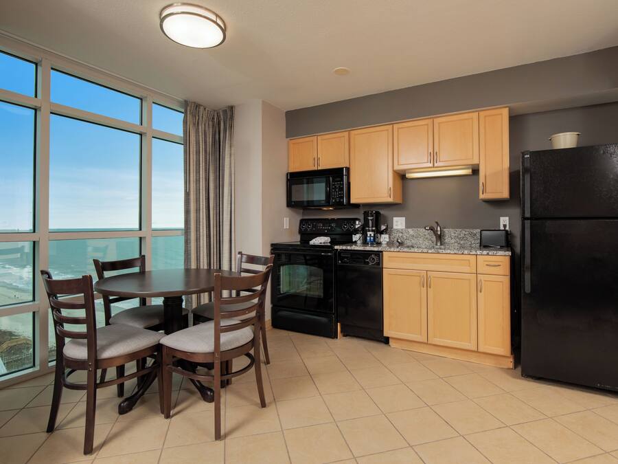 A modern kitchen with wooden cabinets, a black refrigerator, a microwave, and a coffee maker. There is a dining table with chairs and a large window offering a view.