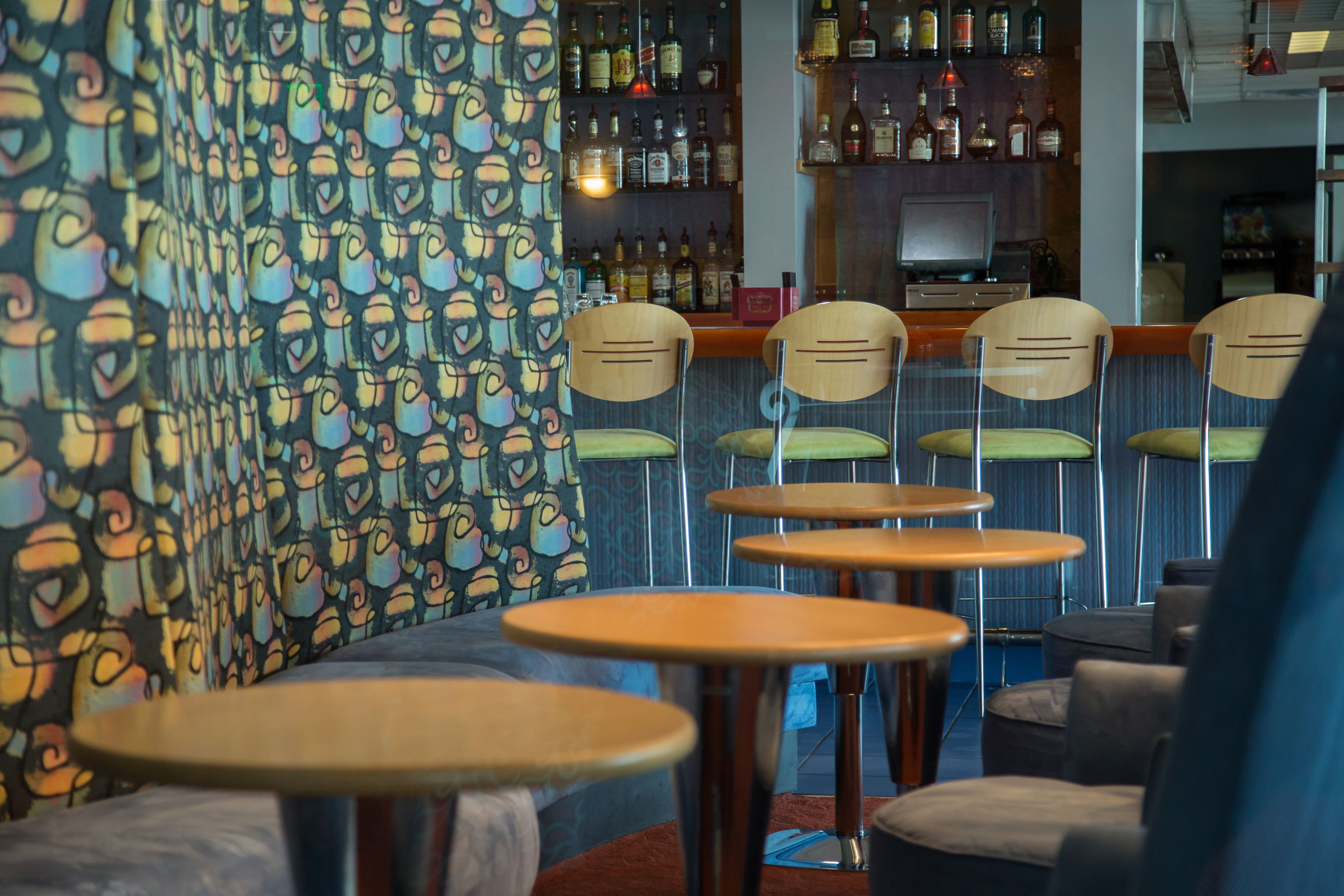 A bar area with multiple wooden tables and chairs, a patterned wall, and shelves with bottles.