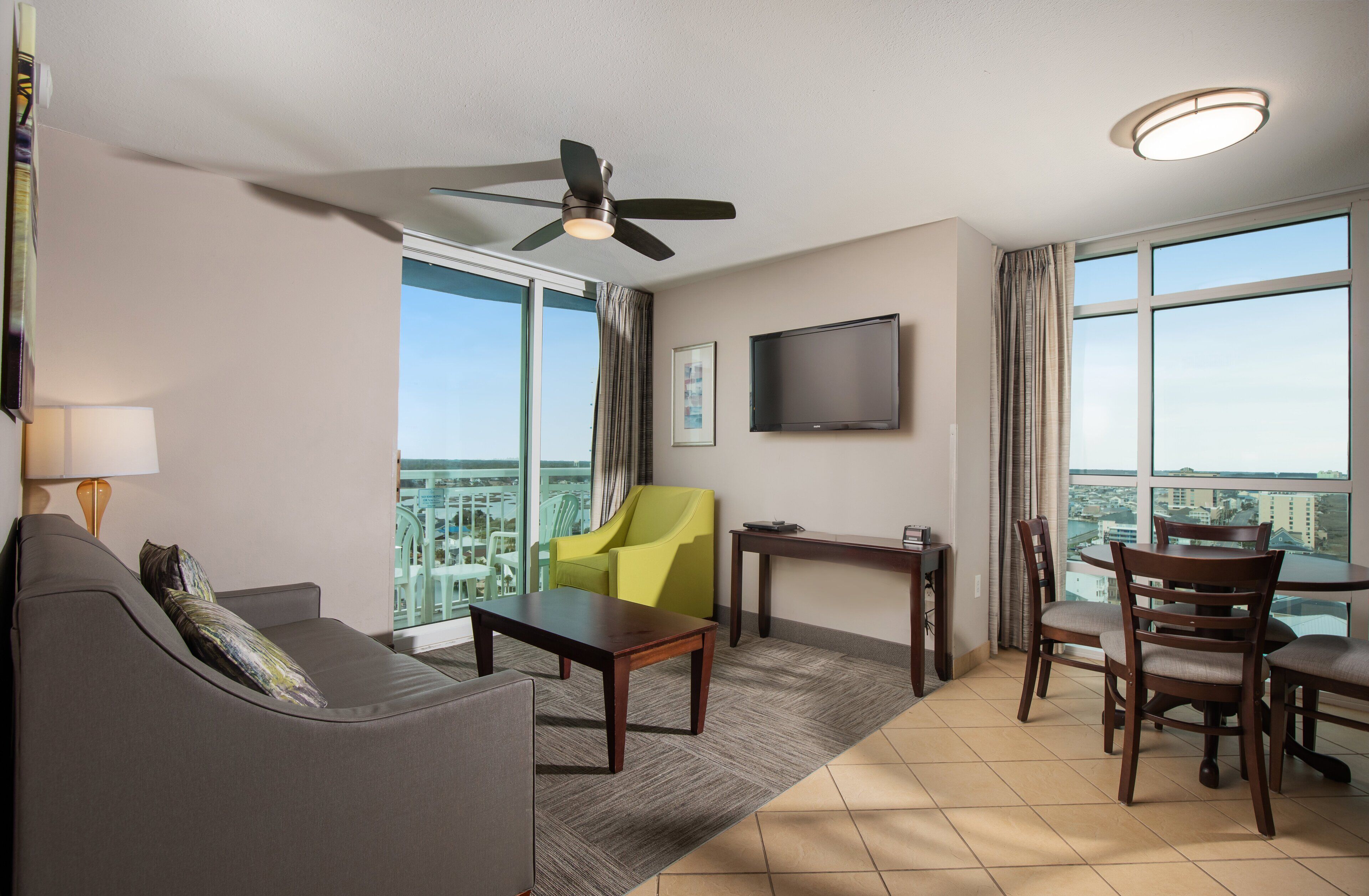 Ein Hotelzimmer mit einer Couch, einem gelben Sessel, einem Esstisch mit Stühlen, einem Deckenventilator, einem Flachbildfernseher und großen Fenstern mit Blick auf die Stadt.