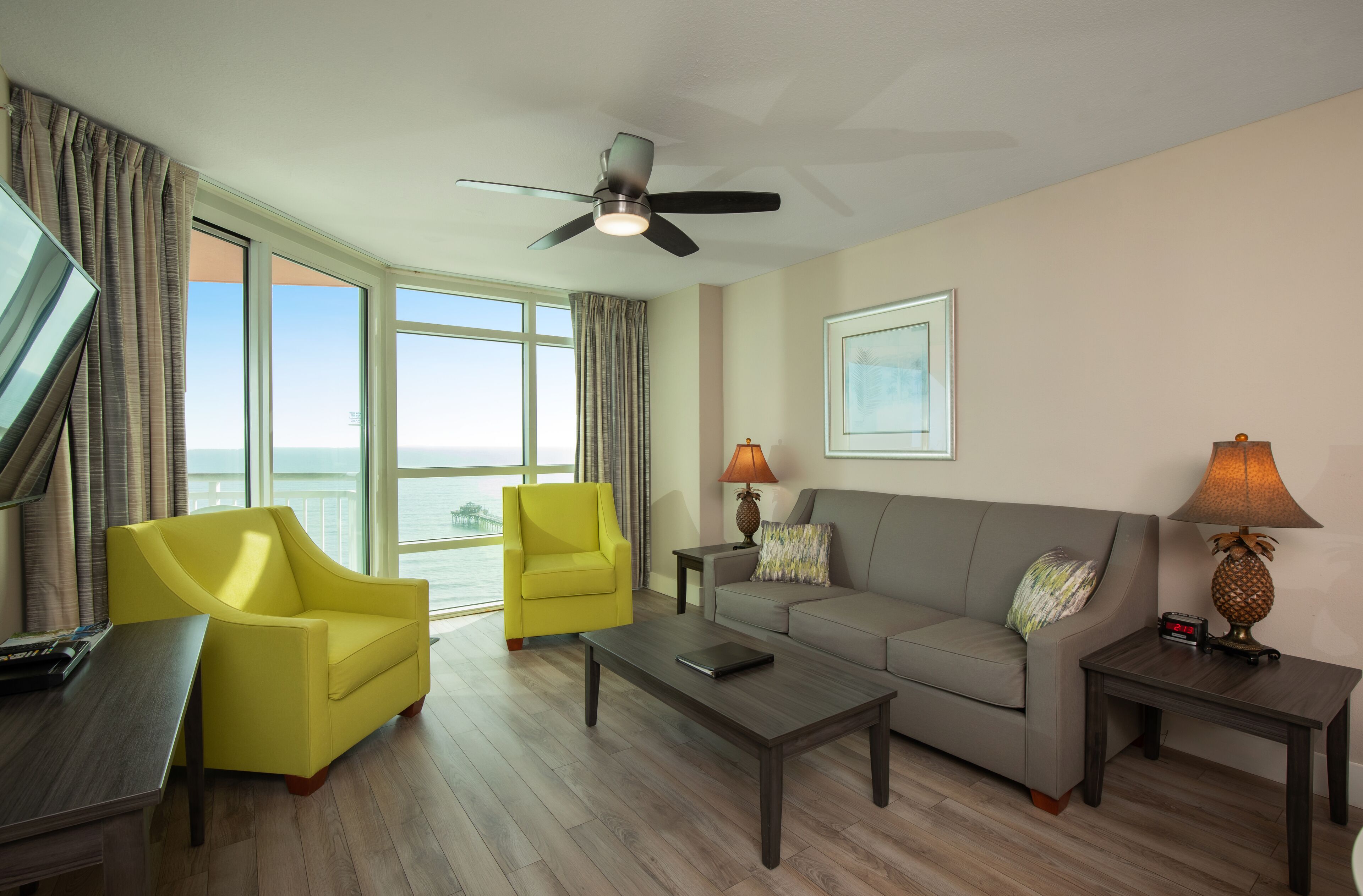 Una sala de estar moderna con un sillón amarillo, un sof á gris y una mesa de centro. Hay un ventilador de techo, un televisor y una lámpara en una mesita auxiliar.