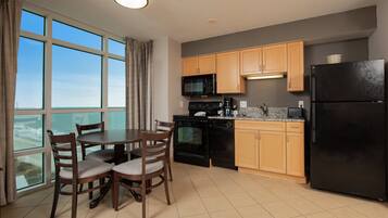 A modern kitchen with wooden cabinets, a black refrigerator, a microwave, and a dishwasher. There is a dining table with chairs and a window with curtains.