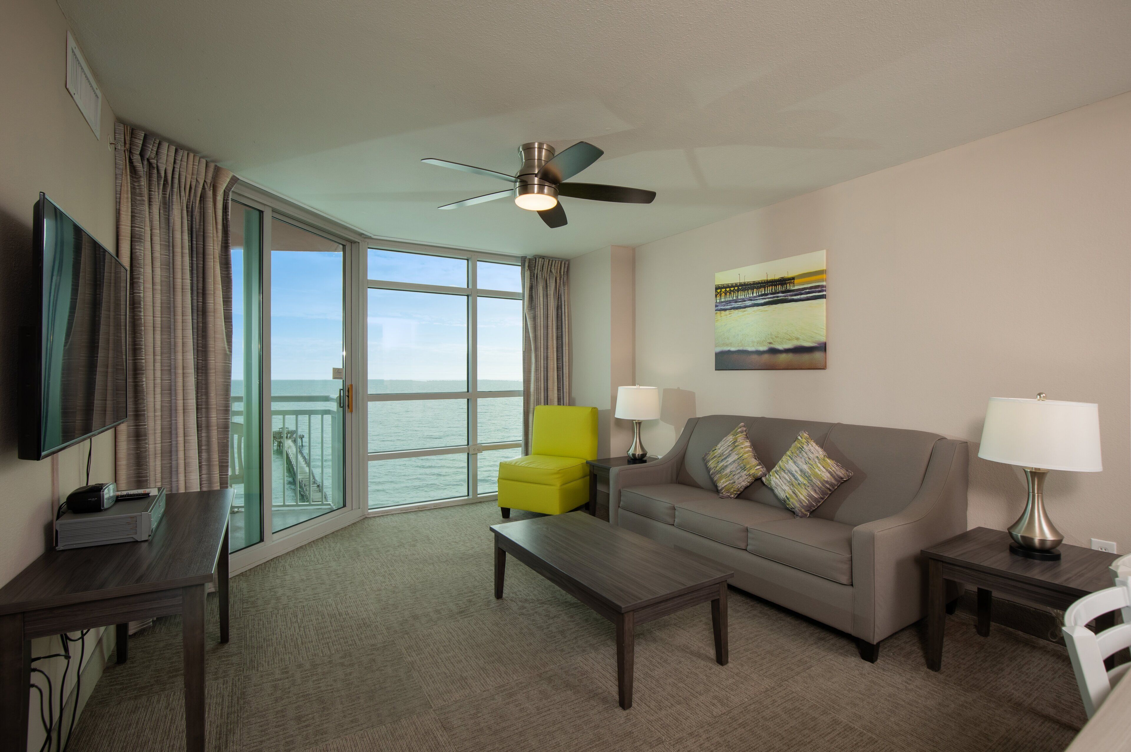 A hotel room with a large window, a sofa, a coffee table, a TV, a lamp, and a ceiling fan.