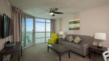 A hotel room with a large window, a sofa, a coffee table, a TV, a lamp, and a ceiling fan.
