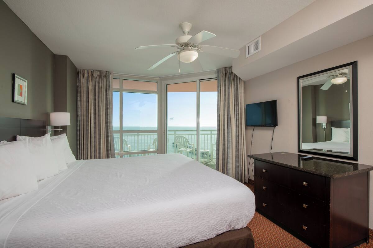 A hotel room with a large bed, a ceiling fan, a dresser, a TV, and a balcony with a view of the ocean.