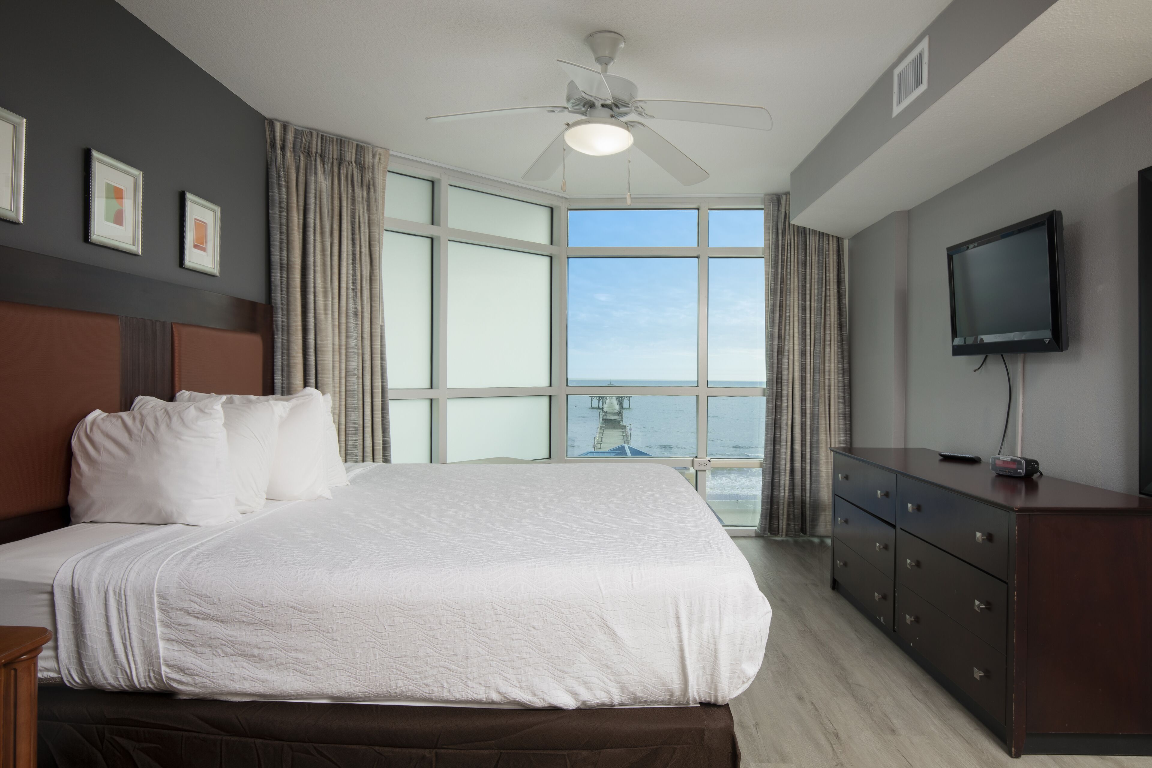 A hotel room with a large bed, a ceiling fan, a TV, and a view of the ocean.