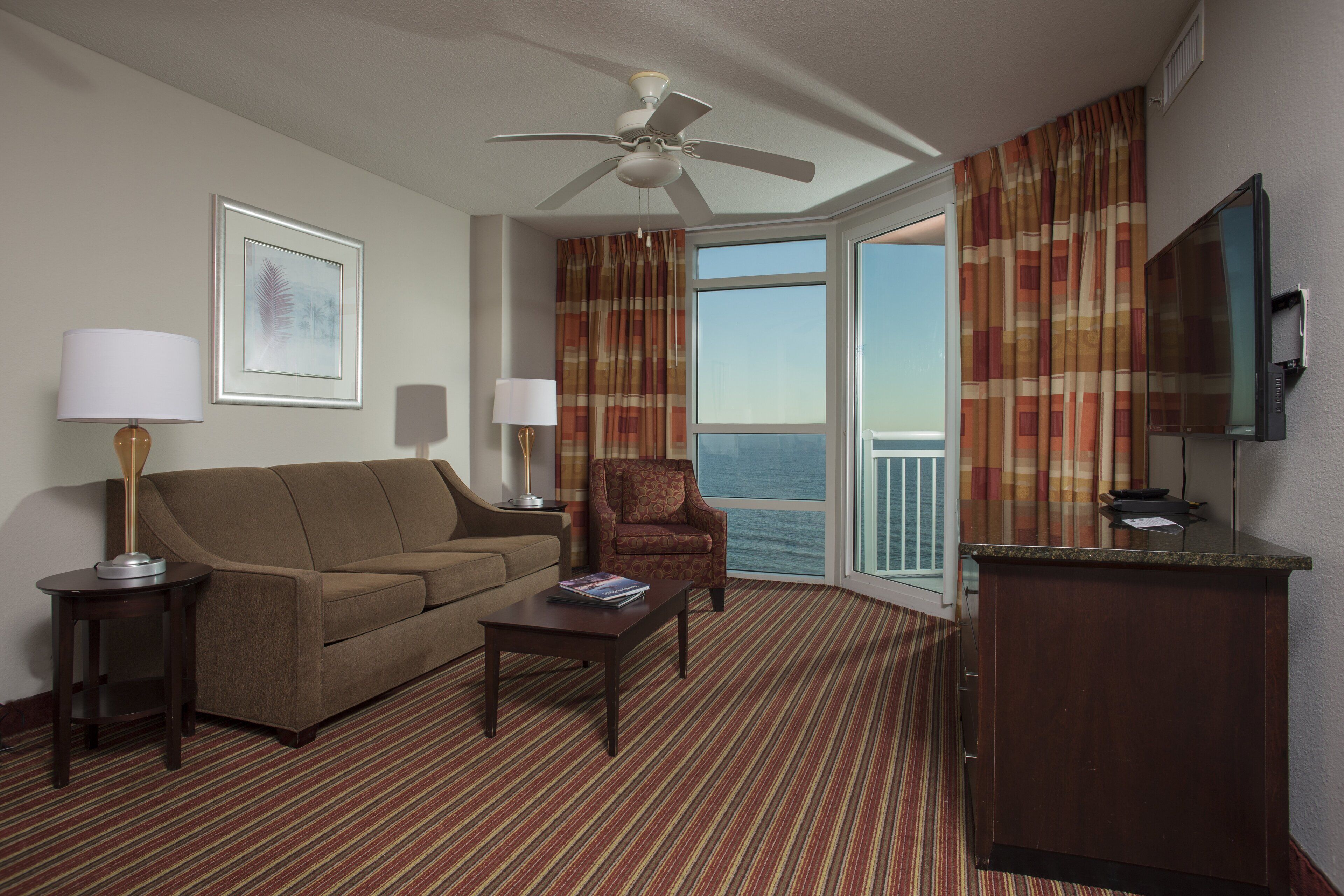 A hotel room with a sofa, armchair, coffee table, TV, and a balcony with curtains.