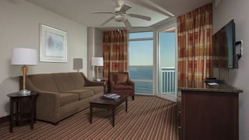 A hotel room with a sofa, armchair, coffee table, TV, and a balcony with curtains.