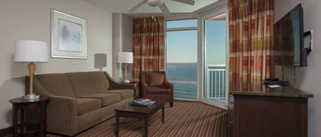 A hotel room with a sofa, armchair, coffee table, TV, and a balcony with curtains.