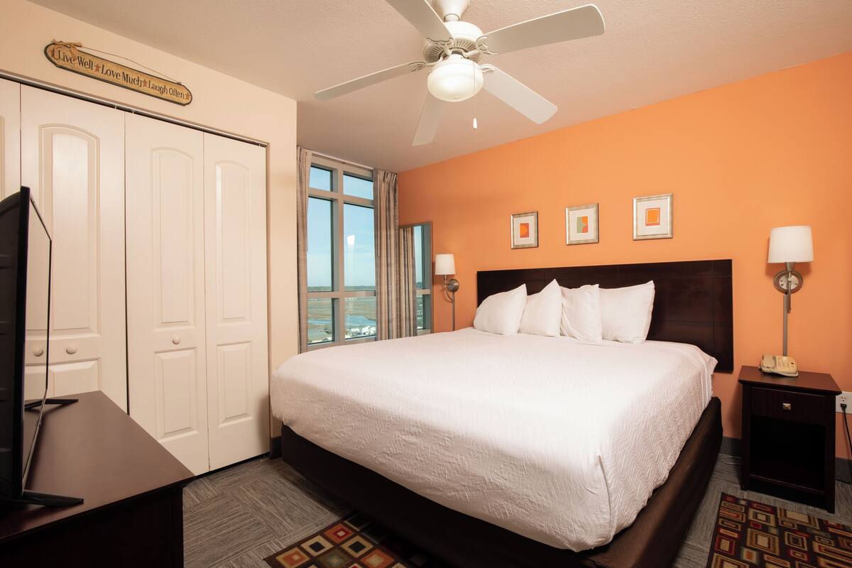 A hotel room with a large bed, a ceiling fan, a TV, and a view of the outside.