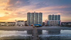 Un hotel frente al agua con un muelle que conduce a la playa, con varios edificios altos y una torre de agua.
