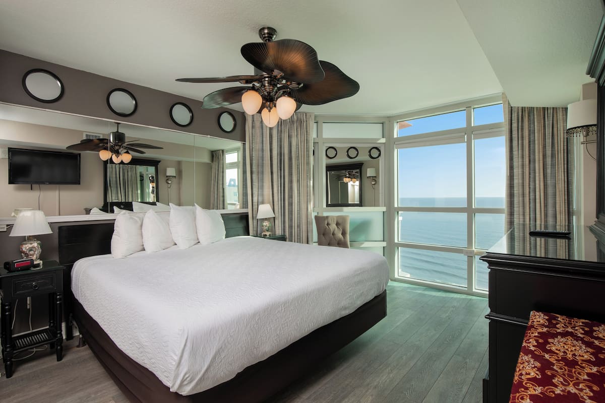 A hotel room with a large bed, a ceiling fan, a TV, and a view of the ocean.
